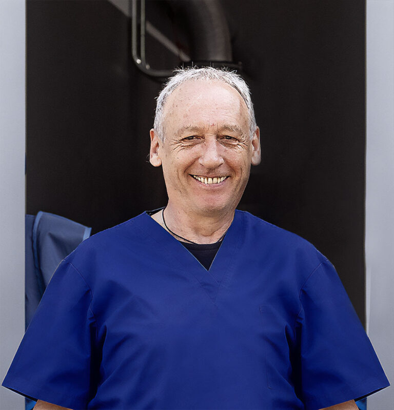 Porträt eines lächelnden Mannes in blauer medizinischer Kleidung. Er hat kurzes, graues Haar und steht vor einem dunklen Hintergrund.