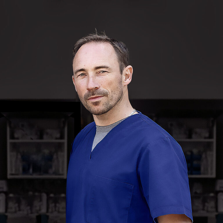 Porträt eines Mannes in blauer medizinischer Kleidung. Er hat kurzes, dunkles Haar und einen leichten Bart. Der Hintergrund ist dunkel und hebt ihn klar hervor.