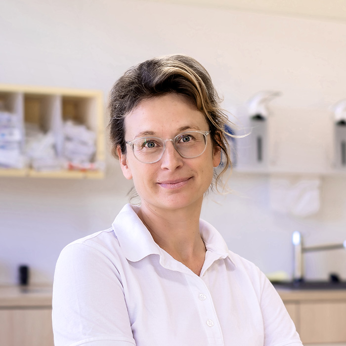 Das Bild zeigt eine Person, die freundlich in die Kamera lächelt. Sie trägt eine weißes Shirt mit Kragen und hat eine entspannte, selbstbewusste Körperhaltung. Die Person hat hellbraunes, leicht hochgestecktes Haar und trägt eine transparente Brille. Im Hintergrund befinden sich unscharf erkennbare Elemente einer Praxisumgebung, wie ein Regal mit aufgeräumten Gegenständen und medizinischem Zubehör. Es handelt sich um Lena Tietje, die Praxisinhaberin der Tierarztpraxis.