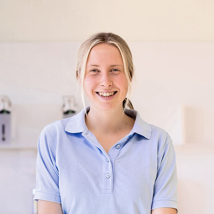 Das Bild zeigt eine junge Frau mit blondem Haar, das zu einem lockeren Pferdeschwanz gebunden ist. Sie trägt ein hellblaues Poloshirt und lächelt freundlich in die Kamera. Ihre Haltung wirkt entspannt und offen. Im Hintergrund ist eine unscharfe Praxisumgebung zu erkennen, mit medizinischen Geräten oder Hygieneartikeln. Die Person auf dem Bild ist Luise Masch, die sich in der Ausbildung zur Tiermedizinischen Fachangestellten (TFA) befindet.