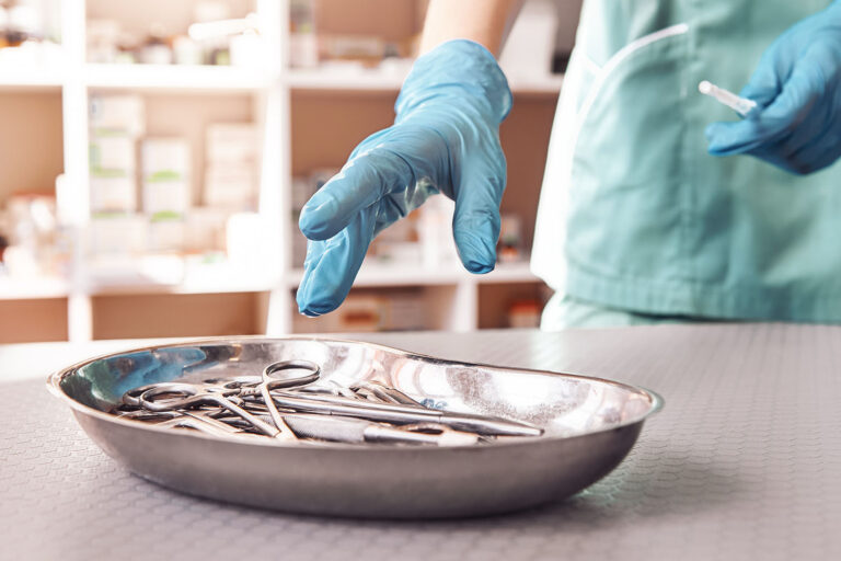 Ein medizinischer Fachmann in grüner OP-Kleidung und blauen Handschuhen greift nach chirurgischen Instrumenten in einer Nierenschale. Im Hintergrund sind Regale mit medizinischen Vorräten zu sehen.
